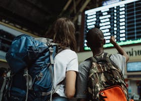 Two backpackers checking the digital board at a train station for travel information.